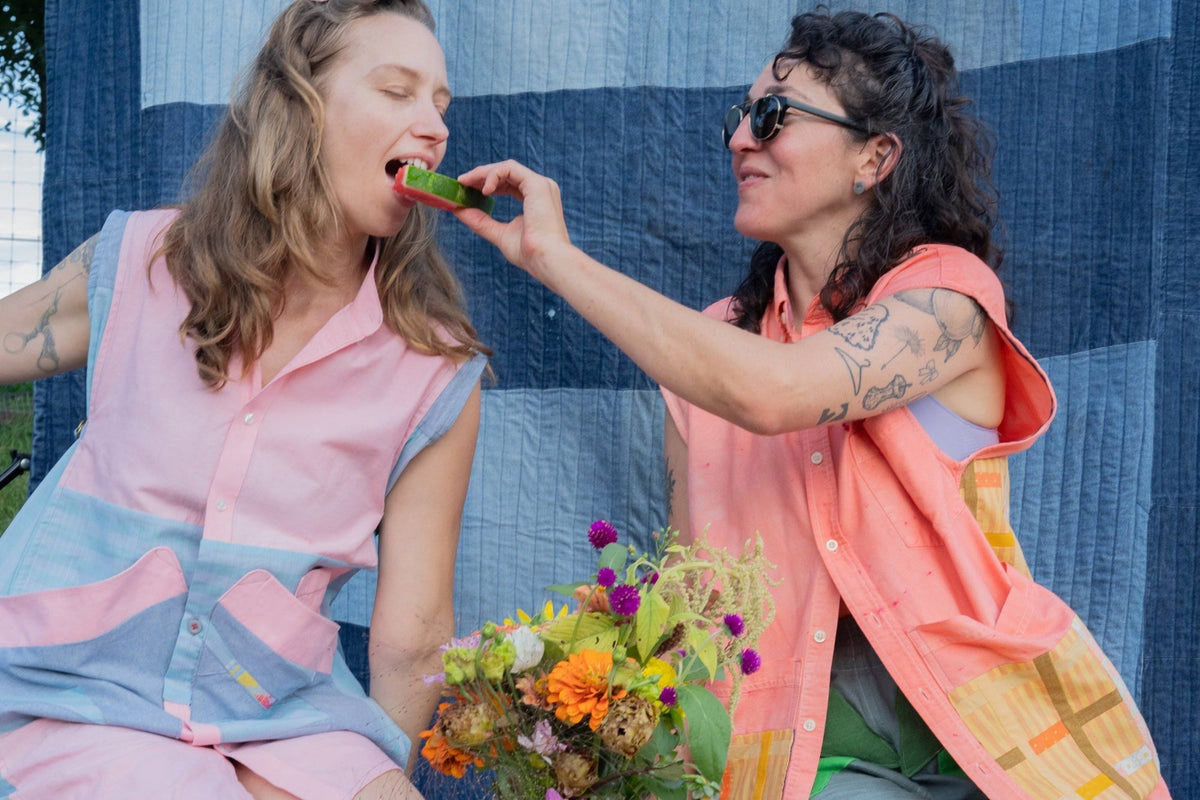 Two women sitting outdoors in front of a blue quilted backdrop, wearing matching pink and blue patchwork button-up shirts. One woman is feeding a green vegetable to the other, with a colorful bouquet of flowers in the foreground.